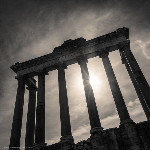 
In a photograph, you can see an old structure standing tall amidst the bright sunlight. The structure seems ancient, with intricate details visible despite the absence of color. It stands in the Roman Forum, a historic site in Rome, Italy. The sunlight casts a stark contrast, outlining the silhouette of the ruin against the sky.