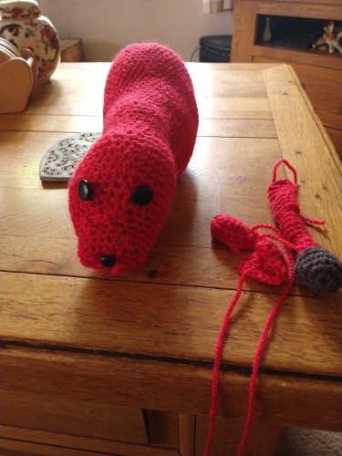 Red crochet shapes on the left a long stuffed shape with two black eyes and black nose. On the right are two small ears and a tail with a brown tip, they will be put together to make a Lion, with legs and mane 