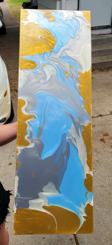 A rectangular canvas being held. the canvas is painted with a strip if bright blue down the center, with white, silver, and gold stretches within.