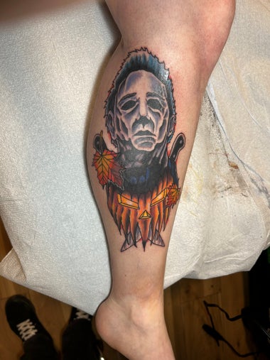 A tattoo of Michael Myers in the Halloween franchise. His neck and collar fade in to a jack-o’-lantern and he is adorned by two butcher knives and two fall leaves