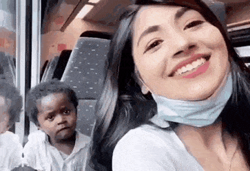 A young woman riding a train takes a video selfie of herself making funny faces as a child in the seat behind her watches and laughs with her.