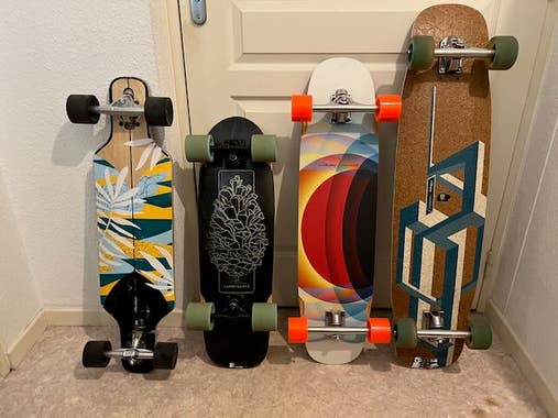 4 longboard standing against a wall and a closed door. Various models, shapes and sizes