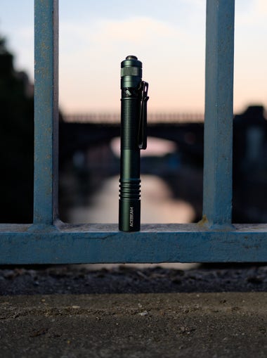 An Acebeam Pokelit 2AA flashilght standing vertically with a canal and bridges in the background