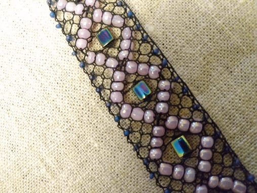 A bracelet about 1 inch wide, with black silk bobbin lace stitches, pink large beads in a diamond shape, and square center beads. Tiny purple seed beads run down the outer edge.