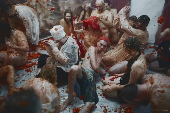Image Transcription:  [ A chaotic scene unravels as a dozen or more people sit, kneel, and lay down in a room that has been waterproofed with clear plastic tarp over a white and deep-red background. Tomatoes, tomato skins, and tomato pulp fly freely in every direction as attendees attempt to cover their eyes or flinch as tomato flies into their faces. Simultaneously, many try to fling tomatoes back. Most appear to be laughing with glee, though some appear to be taking their facefulls of tomato with a calm resignation. Everyone’s white clothes are tinged slightly orange from the pulp as the cloth clings wetly to their skin, their hair matted to their heads-- There is hardly a single inch of the scene that does not contain some smear of tomato innards or a sticky tomato seed or two. Highlights of the image include a man on the right wearing a tall chef’s hat hands upraised to shield his face as he twists away from us. A skinny young man wearing a red bandana on his head lounges with his bony legs facing us at the center of the picture, his face twisted into a puckered grin. Beside him kneels a young woman wearing a black halter top, her face obscured by wet locks of dark messy hair. Towards the back, a woman wearing a very large floppy red hat appears to be having tomato innards smooshed into her colorful headgear by a man who might be entirely nude, though the details are obscured by the chaos of the scene and the orangey, fleshy tint of everyone’s formerly pristine white clothing. ]
