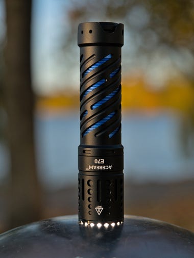 An Acebeam E70 flashlight stands lens-down on a metal ball in front of a blurred lake. Some light escapes around its bezel.