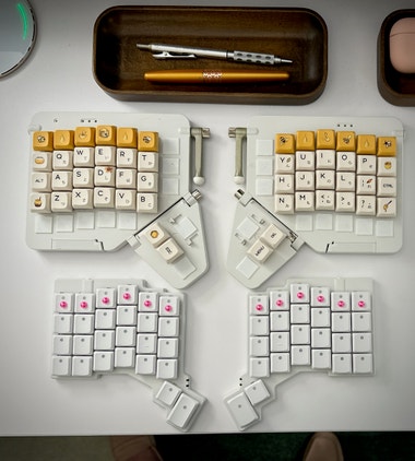 Two split keyboards on a white desk. The upper board is a Moonlander wearing bee keycaps, and is significantly larger than the Voyager board below, which has small raised pink sticker dots on the keycaps of the uppermost row.