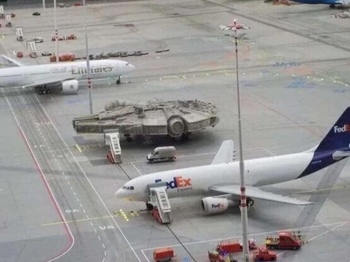 The Millennium Falcon parked on an airfield beside a FedEx airliner.  An Emirates Airliner can be seen beyond. 
