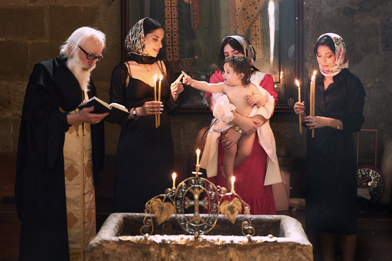 This is what appears to be a formal baptism or christening. A priest, three women, and an infant are gathered around a baptismal font. The priest is on the left holding a Bible; the women all have dark hair and wear scarves and the ones on the outside are holding lit candles. The woman in the middle is the mother and is holding the dark-haired child who is nude, and who is touching fingers with the woman to the left. This looks to be in a cathedral or formal chapel of some kind.