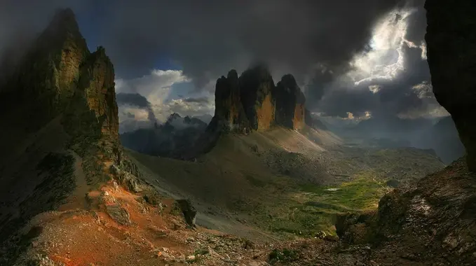 [ A landscape photo featuring tall mountains jutting out from a rolling landscape under a darkly-lit sky, save for one shock of sunlight in the upper right corner where dark clouds have parted ]