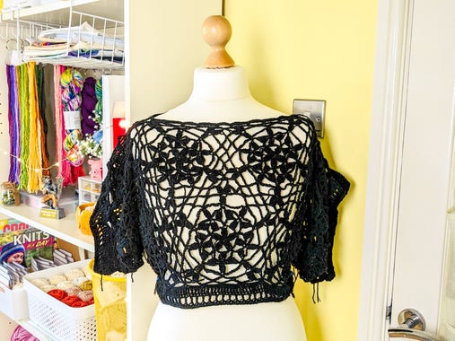 A white dressmakers dummy stands in a yellow room. Behind it are white shelves holding all manner of bright and colourful craft supplies. The dummy is wearing a very open, lacy black crochet crop top with short sleeves. The top is made up of a repeating, vaguely floral motif, with an eyelet border at the bottom and around the sleeves.
