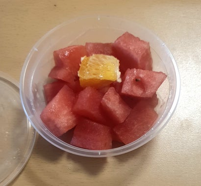 A 350ml or 12oz plastic round container sold as a fruit salad. It contains 1 inch chunks of chopped fruit. All chunks are watermelon except a single chunk of orange carefully placed on top.