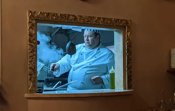 [ What initially appears to be a painting of a rotund chef in a traditional uniform, mounted in an ornate gold frame and hanging on the wall is-- Upon closer inspection-- a window into a real life kitchen. The optical illusion is helped somewhat by the stark difference in lighting; the chef and the kitchen are under cool-toned florescent or LED lights, while the room where we sit is illuminated in soft warm lighting with darker stucco walls, creating the illusion that the kitchen scene is a sun-faded painting. The chef himself wears a double-breasted white chef’s jacket with sleeves that are deeply creased from being pushed up habitually. The buttons strain against his considerable bulk and the toque sits on his brow snugly. His pale face is creased in concentration as he tips a fry basket into a pan and a cloud of steam billows upwards. A clear squeeze bottle of oil sits at his elbow and various metal handles and kitchen appliances clutter the sleek industrial kitchen. The bottom of the “frame” is a shelf, suggesting that this is less of a fine dining situation and more of a fast-casual restaurant where management installed the window to get food out as fast as possible to customers. ]