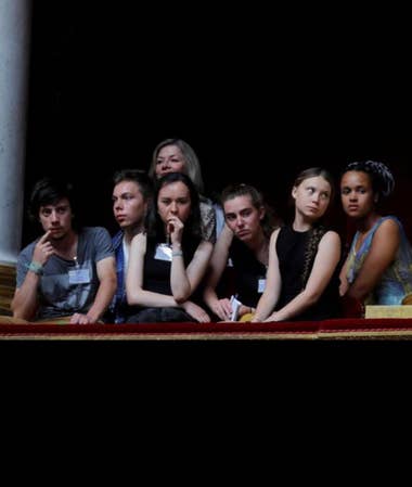 [ Six young adults sit behind a plush red velvet railing, clothed in shades of blue and black with their bare arms folded, touching their faces, or resting in their laps.  Their gazes are focused outward and their expressions are careworn and concerned.  One older woman peeks from from behind the group, she is the only one with any trace of a smile on her partially obscured face.  Most recognizable among them is climate activist Greta Thunberg, whose eyes are gazing up and away from the rest of the group-- It is unclear if something grabbed her attention, or the photograph was taken mid-eyeroll.  The background is entirely black, blending into their dark clothing, making their faces and arms appear luminous as they emerge from the shadows.  The scene is one of quiet tension and profound disappointment.  ]