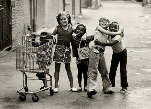 A black-and-white photo of five children, three with black skin and two with white skin, hanging out in the street. The youngest child is pushing a shopping cart.