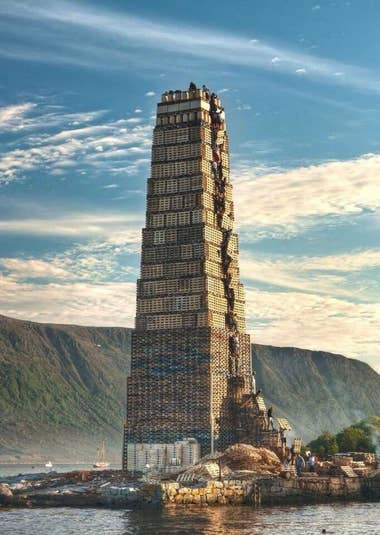 Image of a giant tower made up of crates, being constructed by people. It's on a small island in the Netherlands and resembles scenes depicting the Tower of Babel in the Bible.