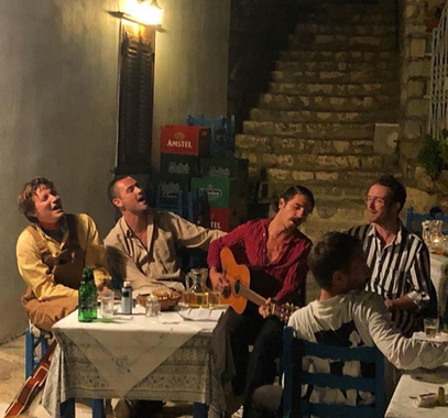 This photo is in an ancient looking alleyway in Europe. Four men are seated behind a small table with wine and bread upon it. The man in the middle is playing a guitar; the other men are looking on and singing. There is an old stone stairway behind the group, and a whitewashed wall with a wooden window to the left. The men are in contemporary clothing. A fifth man is facing away from the viewer, looking to his right.