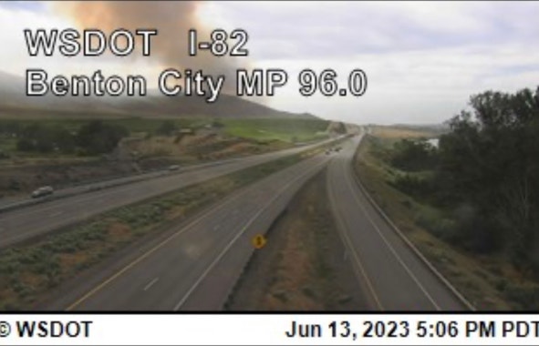 A rural freeway with a smoke plume rising behind a hill in the distance just left of center. Text overlayed over the image says WSDOT I-82 Benton City MP 96.0 and the time stamp is 17:06 June 13, 2023