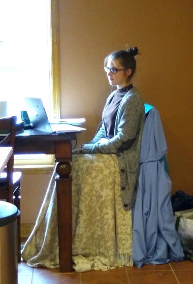 Image Transcription:  [ A pale woman with a tight hair bun and glasses sits at a dining room table, her hands folded in her lap, with a laptop open in front of her. The bright sunlight from the window immediately to the left bathes her in the cold sunlight of early morning. On the table in front of her, difficult to make out in the over-exposed area of the photo, are a can of soda and a notebook. She is dressed in an oversized pale green knitted cardigan and a dark purple turtleneck shirt, and what appears at first to be an exquisitely decorated yellow brocade skirt with fringe trim is, upon closer inspection, a blanket she has draped over her lap for warmth. It is simply an optical illusion that she is wearing some kind of grandiose ballgown; An illusion heightened by a long light blue jacket hung over the chair she is sitting on that looks something like a bustle. The room she is sitting in has warm-hued walls and a tile floor to match, giving it a vaguely historic vibe. ]