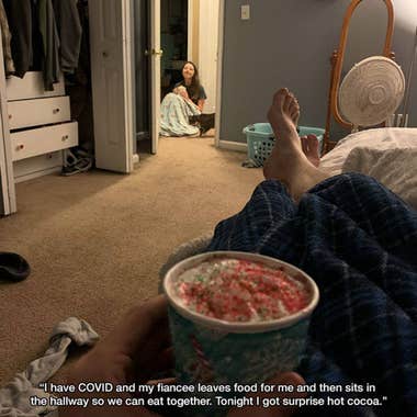 A photo from the perspective of a man lying down in bed holding a small cup of cocoa in the foreground. In the background, a woman sits looking at the camera with a loving expression, holding a cup of something. A cat sniffs the blanket that covers her legs. Caption: "I have COVID and my fiancee leaves food for me and then sits in the hallway so we can eat together. Tonight I got surprise hot cocoa."