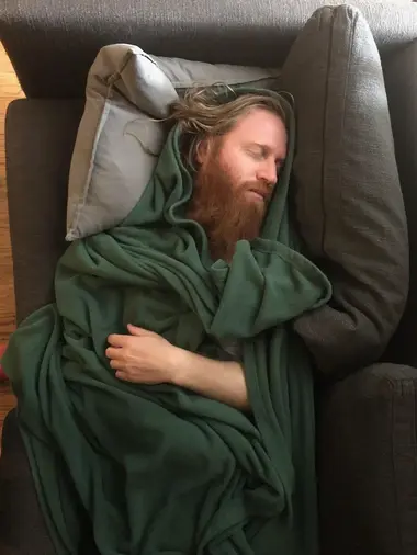 Image Transcription:  [ A young and handsome man, with a reddish beard and long blond hair sleeps on a couch, wrapped in a green blanket. At first the photo appears as if he is standing with his eyes closed, saint-like, but closer inspection reveals that is is in fact napping on a grey sofa and the photographer snapped this photo by holding the camera above him. His form is almost entirely obscured by the deep folds of the green blanket, the only parts of the man which are visible is his face, a few fly-away strands of long blond hair escaping the place where the blanket has formed a hood, his reddish beard, and his bent left arm which rests in a loose fist on his chest. The lighting is soft and the scene feels vaguely religious, reminiscent of paintings showing a dead Christ before resurrection. ]