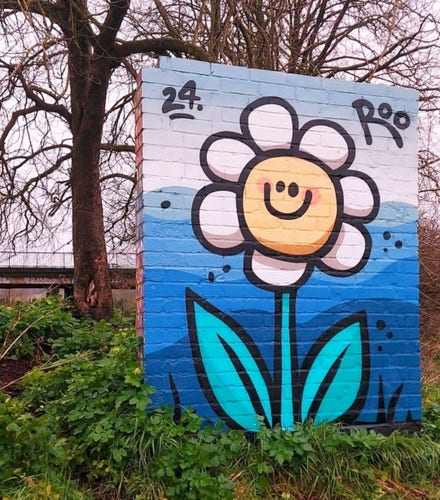 Streetartwall. The cheerful mural of a smiling flower has been sprayed on a small free-standing section of wall. The cheerful yellow flower with white petals has a smiley face, turquoise leaves and stems and stands in front of a hilly landscape of varying shades of blue. The name of the artist ROO and the year are also noted. (The photo shows grass and clover around the piece of wall, with brown trees and a bridge railing behind it)