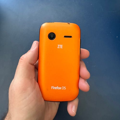 A hand holding an orange device, showing the back. “FirefoxOS” branding is visible, indicating this is very much not a Rabbit. 