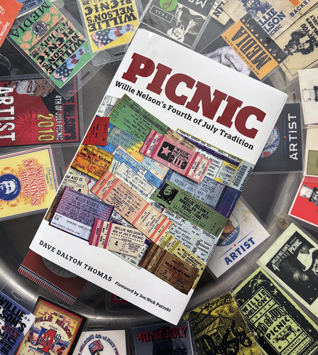 Photo of Dave Dalton Thomas' new coffee-table book entitled "Picnic: Willie Nelson's Fourth of July Tradition". The book is featured on a custom-made coffee table featuring a collection of laminated media lanyards in-laid into the glass of the table. On top in the middle of the table is the book with its front cover featuring a collage of 4th of July Picnic concert ticket stubs in assorted colors - it's a fitting pastiche for such a long tradition. 