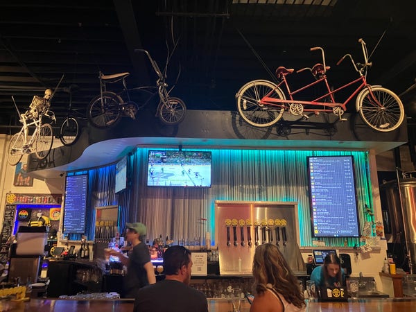 A photo of a bar with some patrons and bartenders tending to them. Three bicycles hang above the bar, one of them is a tandem and one is being ridden by a plastic skeleton.