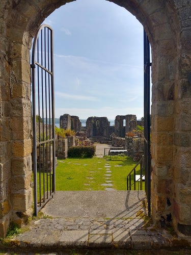 à travers un portail ouvert, on voit les ruines du chœur d'une ancienne église, le sol est couvert d'herbe, au loin, on aperçoit la mer, le ciel est bleu.