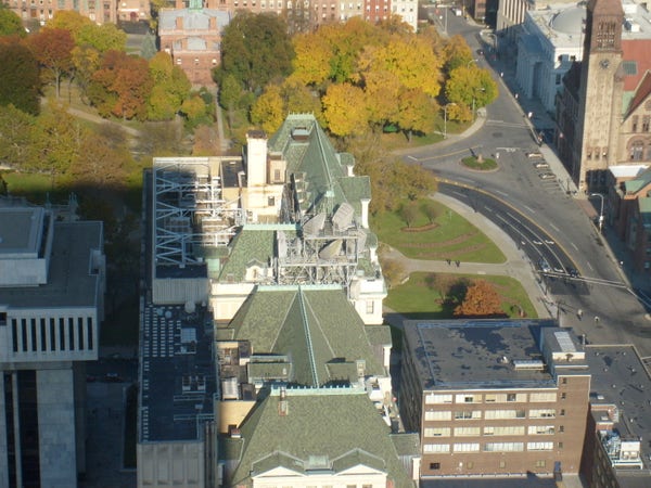 Photo taken from several hundred feet in the air looking down at the ground and the roofs of several multi-story buildings. The building at center has a greenish colored roof with a complex collection of HVAC equipment and several large antennas mounted to it.