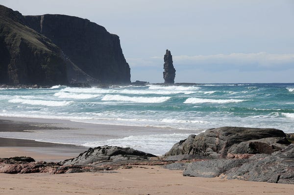 Sandwood Bay, south of Cape Wrath in Sutherland. The image shows large waves rolling in from the right over a light blue sea, with sand and rounded rocks in the foreground. Intruding into the image from the left are black cliffs. These end in the middle of the picture and then there is an elegant sea stack.
