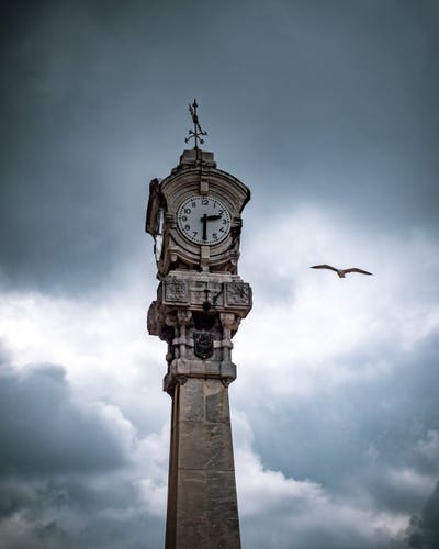 A clock on top of a marble pillar with dramatic clouds in the background. On the right side of the clock a seagull fly towards the clock