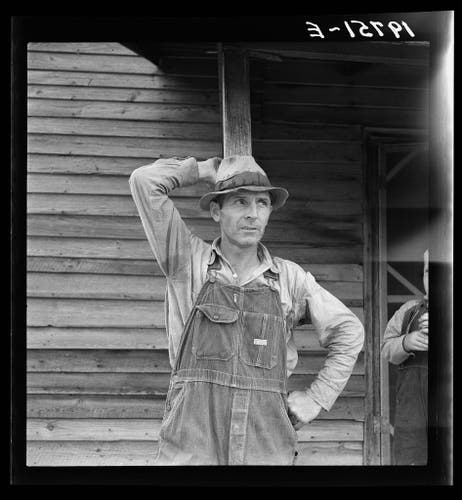  The image is a vintage black and white photograph capturing a moment in the life of an individual, likely from the mid-20th century. It shows a person wearing a wide-brimmed hat and overalls, standing with their hand on the side of their head. They appear to be resting or contemplating, as they lean against what looks like a wooden structure, possibly related to agriculture or farm work. The setting is outdoors, under a clear sky. There's an additional text overlay that reads "Tobacco sharecropper tells about his prospects. Person County, North Carolina," which suggests the photo might be part of a documentary or journalistic project focused on the life and times of sharecroppers in that region. The image provides a glimpse into the daily experiences and environment of this individual's occupation, offering insight into their working conditions and community.