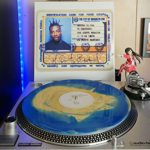 A Yellow & Blue swirl vinyl record sits on a turntable. Behind the turntable, a vinyl album outer sleeve is displayed. The front cover shows an ID card featuring Ol' Dirty Bastard's image. 
