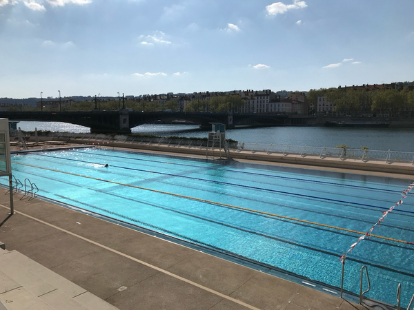 Au premier plan, le bassin olympique de la Piscine du Rhône à Lyon. Une seule personne nage seule dans cet immense bassin. Au second plan le Rhône, le pont, puis le quai et les immeubles de la presqu’île. Le soleil est bleu ciel, la piscine est bleue piscine et le Rhône est bleu gris. 