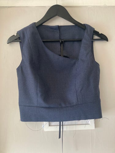 Bodice of a dress on a hanger. Fabric is blue/black microcheck: almost plain. It’s a sleeveless dress with princess seams and a waistband. The neckline is asymmetrical, with a  long diagonal sweep from one shoulder to just above the bust, and a much narrower shoulder strap on the other side