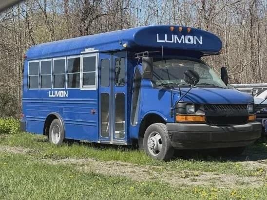A small blue Lumon Industries bus parked in some grass.