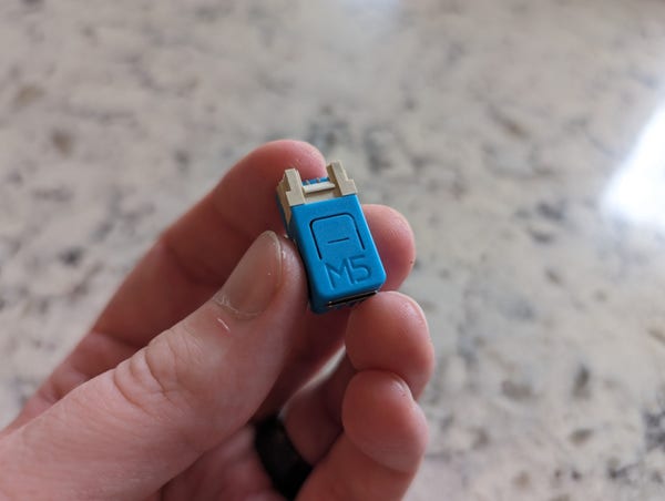 Hand holding M5STACK C6Nano dev board. Small blue enclosure, approximately 1cmX2cm. There is a USB c port on one end, and a Grove connector on the other.