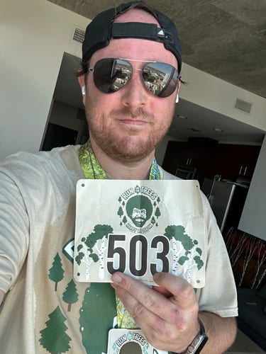 Selfie of me, still sweaty, sunglasses on, wearing a beige “Run for the Trees” shirt featuring Bob Ross’ face. Holding my bib number, 503.  