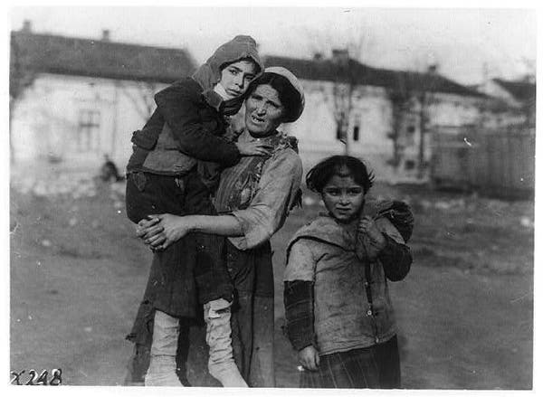 In this black and white photograph, a mother is seen walking with two of her children on a street. The mother is dressed in traditional attire, suggesting that the photo might have been taken during a time when such clothing was common. She appears to be carrying one child while the other walks alongside them, holding onto their hand. There are buildings and trees visible in the background, indicating an urban setting with some greenery around. The image captures a moment of everyday life, conveying a sense of familial bonding amidst a bustling environment.