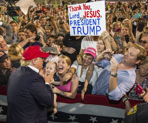 Trump greets a crowd of rapturous white people. One holds up a baby. Another holds a sign that reads "Thank You Lord Jesus, for President Trump"