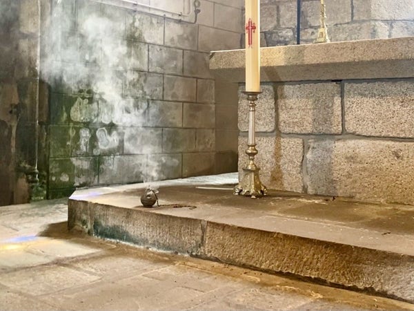A church interior corner with a large liturgical candle on a stand, incense smoking on a stone step, mossy wall. 