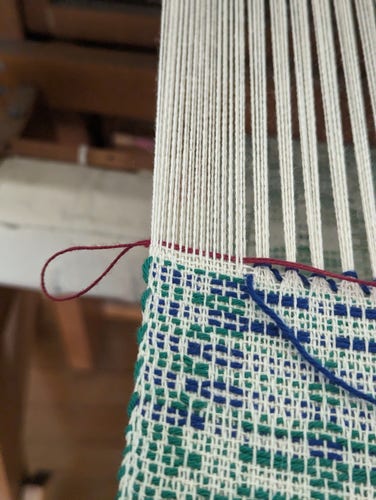 the end of a piece of weaving, still on the loom. a short piece of red yarn is woven across the last inch of warp threads, with a loop sticking out at the edge of the cloth.