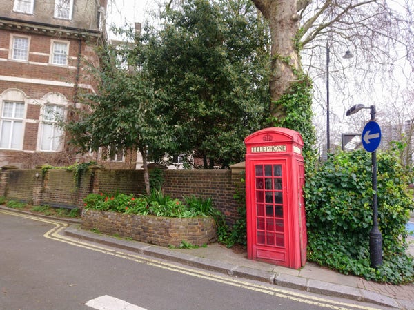 London street corner with an old-fashioned red telephone box surrounded by plants and trees against a red brick wall. 