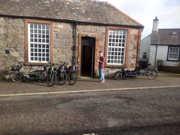 Some of the village hire bikes outside the village hall. The hall is solidly built of grey stone with large windows and a slate roof. Outside are two cargo bikes, and three conventional ebikes.