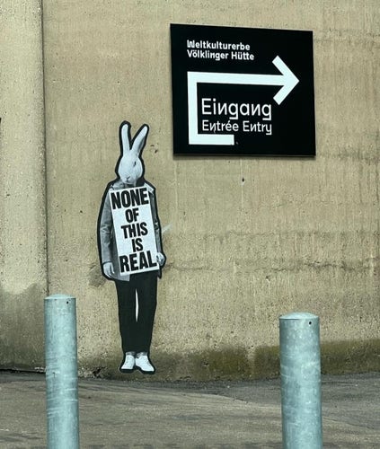 Streetartwall. An upright rabbit with a sign has been painted and glued onto a beige concrete wall. He is wearing black trousers, sports shoes and a gray jacket. He is standing next to the entrance to an exhibition space for urban art. The sign reads: "None of this is real". Next to it is a painted sign with an arrow, the word Eingang/Entry/Entree and World Cultural Heritage "Völklinger Hütte".
Info: The "Urban Art Biennale" is the largest exhibition of urban art in Europe. Since 2011, artists from all over the world have been given the opportunity to present their works in a former ironworks, which is now a UNESCO World Heritage. 
Opening April 28 / 3 pm.  Admission is free on this day from 2 pm. Access to the old steelworks is only partially barrier-free.