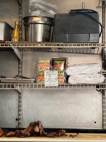 Metal shelving in a food cart kitchen with various items including cans of SPAM with a sign posted in front reading "NOT 4 CARLOS” with a frowny face, a pot, a bottle, and charred food on the bottom shelf.