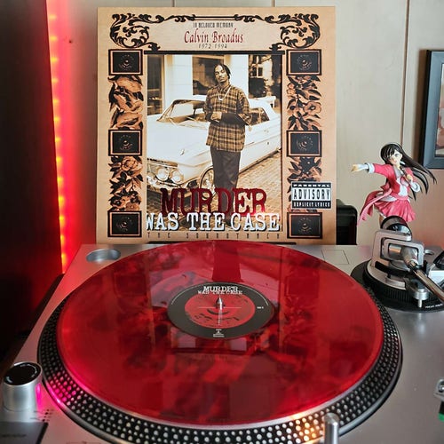 A translucent red vinyl record sits on a turntable. Behind the turntable, a vinyl album outer sleeve is displayed. The front cover shows Snoop Dogg standing in front of a lowrider with the surrounding frame looking like an obituary. 