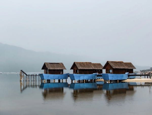 Air is heavy with misty, a cluster of tiny houses, looking like blue boats stand over the still waters of the lagoon, casting beautiful reflections on the water. Scene of a very simple life.